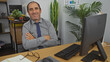 © Krakenimages.com - A smiling hispanic elderly man in an office setting, dressed in business attire, sits comfortably with his arms crossed at a desk with a computer, surrounded by plants and office equipment.