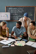 © Seventyfour - Vertical shot of young Black man talking to female teacher sitting at desk in group discussion, while practicing new words in speaking club session at language school for adult learners