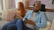 © Krakenimages.com - Interracial couple sitting together on a couch in a cozy living room, both using smartphones, showcasing modern relationships and technology in an indoor home setting