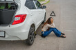 © Odua Images - A woman is experiencing stress as she sits next to her car that has a flat tire on the roadside