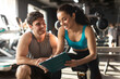 © Home-stock - Professional female personal trainer talking with male client, discussing nutrition or training plan on clipboard in modern gym interior