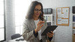 © Krakenimages.com - Woman in office working on tablet, standing near window with brick wall, wearing glasses and checkered blazer, showcasing focused professional environment indoors.