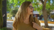 © Krakenimages.com - Woman with blonde hair and glasses standing outdoors, looking to the side in an urban park setting on a sunny day