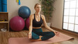 © Krakenimages.com - Woman meditating on yoga mat in gym with exercise balls and plants, wearing sportswear, showcasing serenity and focus in a bright indoor setting.