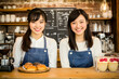 © Putu - Two smiling Asian women in denim aprons stand behind a bakery counter showcasing fresh pastries and desserts.  They offer a friendly and welcoming atmosphere.