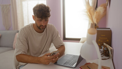 Wall Mural - Young man using phone in living room indoors with laptop on desk and books in the background in a cozy home setting.