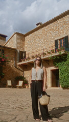 Wall Mural - Young, hispanic, woman enjoying outdoors in historic, stone, building courtyard in mallorca, spain, holding wicker, bag and wearing sunglasses on sunny day