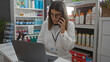© Krakenimages.com - Female pharmacist with brunette hair and glasses talking on the phone while working on a laptop in a well-organized indoor pharmacy setting.