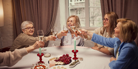 Cozy atmosphere with friends. Elderly women gathering in warm living room a home, drinking wine eating grapes and cheese, cheerful talking. Concept of elderly lifestyle, positive emotions