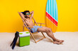 © deagreez - Young man relaxing on beach chair by a colorful surfboard with a yellow background, wearing sunglasses and swim shorts.