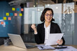 © Liubomir - Joyful businesswoman in office reviews document, celebrates success. Desk displays laptop, papers. Team collaboration shown by colorful sticky notes.