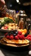 © Michal - A person is pouring maple syrup onto pancakes, which have berries on top and a stack of other pancakes
