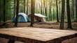 © organik - A empty wooden table in the foreground with a tent and trees in the blurred background, suggesting an outdoor camping scene in a forest