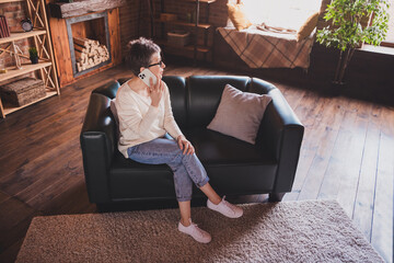 Wall Mural - Elderly woman with short grey hair in casual style pullover sits on a sofa talking on phone in a cozy home interior