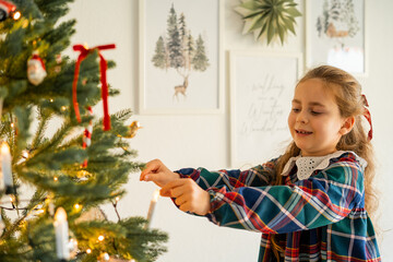 Cute happy little girl decorating Christmas tree at home. Winter holidays.