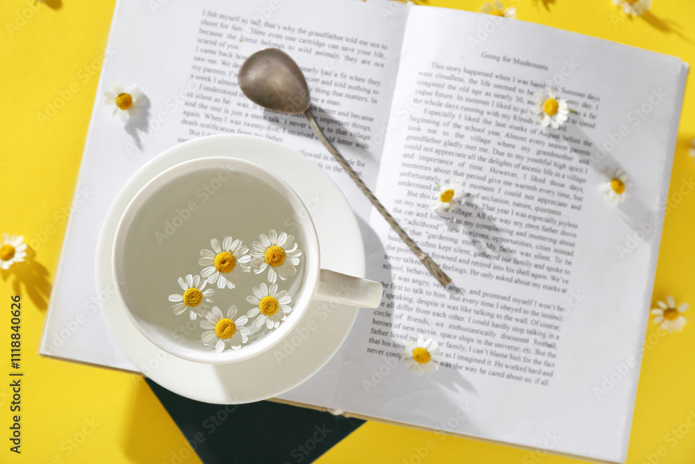 Cup, saucer, tea spoon, book and chamomile flowers on yellow background. Top view