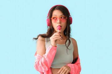 Wall Mural - Beautiful young Asian woman in stylish sunglasses and modern headphones with sweet lollipop on blue background