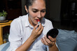 © Marcos - Latin adult Woman applying lipstick and putting on makeup and painting her lips in color red at home in Mexico Latin America