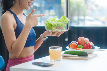 A young Asian housewife enjoys a healthy lifestyle at home, balancing fitness routines with relaxation. She sits on a modern sofa, eating a fresh salad and fruit while embracing healthy habits.