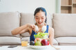 © Phushutter - A young Asian housewife enjoys a healthy lifestyle at home, balancing fitness routines with relaxation. She sits on a modern sofa, eating a fresh salad and fruit while embracing healthy habits.