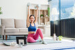 © Phushutter - A young Asian housewife sits on sofa in her living room, using fitness app on her mobile with headphones. dumbbells and whey protein, she balances yoga, workouts, vegetarian diet and weight loss.