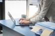 © Phushutter - A young Asian businesswoman works alone at a desk with a laptop, stack of papers, and documents. She looks happy and successful, managing accounts and preparing financial statements in daylight.