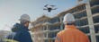 © Wannaeka - Construction workers in hard hats standing in front of building, with drone, scaffolding, and cranes at sunny construction site