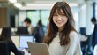 © Ganesha - Smiling Japanese businesswoman standing in an office, holding her laptop. She is wearing casual clothes, and other people working behind her are blurred.,generated ai.