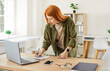 © Studio Romantic - Portrait of a young redhead business woman in casual clothes working with laptop computer in office standing at desk near her workplace and writing something in notepad planning work day.