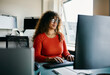 © Flamingo Images - Focused Woman, With a smile on her lips, working on a Computer in Modern Office Environment. She is wearing an orange shirt and glasses.