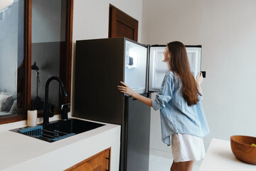 Canvas Print - A young woman happily exploring her fridge in a modern kitchen, dressed casually in a denim shirt and shorts, radiating a sense of joy and wellness