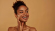 © sajid Pixel - Happy African American woman with bare shoulders applying serum, isolated on beige.
