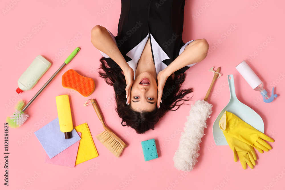 Surprised young housemaid with different cleaning supplies lying on pink background