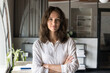 © fizkes - Head shot portrait of pretty young businesswoman posing with arms crossed in modern workspace, looking at camera with serious, peaceful expression. Motivated female business leader, competence, career