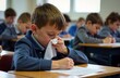 © Kirill - A boy sits at a school desk, the boy is sad and sick, picks his nose with a white handkerchief