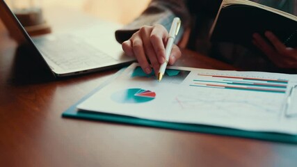 Wall Mural - Businesswoman reviewing financial reports and graphs, highlighting key figures with a pen, and taking notes in a notebook while analyzing data in an office setting