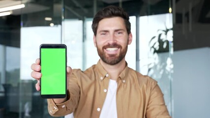 Handsome businessman presenting mobile phone with green screen standing in modern business office. Color key, vertical template layout for advertising. Smiling man looking at camera holding smartphone