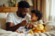 © KateMur - Young Black father giving cold medicine to his child, sitting together on the bed with a comforting atmosphere.