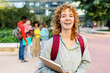 © Xavier Lorenzo - Smiling portrait of ginger student holding a binder on university campus with her friends talking on the background. Education lifestyle and youth concept