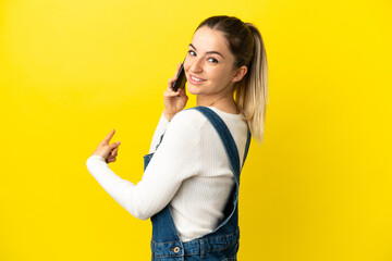 Wall Mural - Young woman using mobile phone over isolated yellow background pointing back