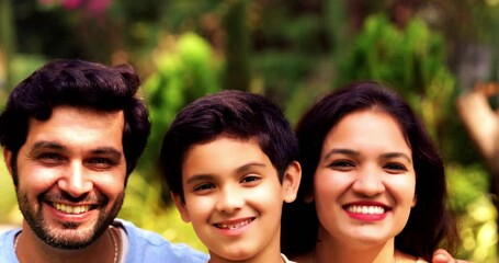 Wall Mural - Indian Happy Family of Four Sitting Together Outdoors on a Lawn in a Park, Kids on Parents’ Laps, Smiling and Enjoying Quality Bonding Time Amidst Nature