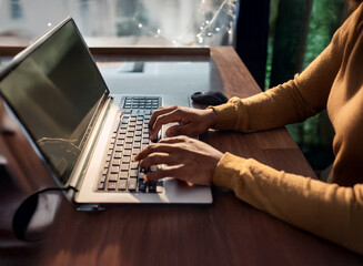 Aufkleber - Close-up of hands typing on a laptop keyboard in a sunlit workspace.  The person is wearing a mustard yellow long-sleeved shirt.  The scene is warm and inviting, suggesting productivity and focus.
