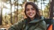© Infinity Lens - Beautiful young woman with brunette hair camping in the forest wilderness, sitting in a camping chair, smiling and looking at the camera. enjoying morning sunshine in the woods, happy female camper.