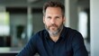 © Formoney - A man with a well-groomed beard is seated at a sleek desk in a contemporary office. Natural light fills the space, highlighting his thoughtful expression and stylish attire