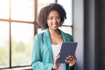 Leinwandbilder - In a bright office environment, a young professional with curly hair holds a tablet while smiling. She stands near a window, reflecting teamwork and innovation in the corporate world.