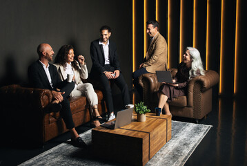 Fototapete - Five professionals engage in a lively discussion in a stylish office space. They share ideas on business strategies while sitting on cozy sofas around a wooden coffee table.