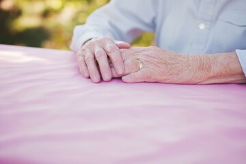 Poster - Elderly hands gently clasping each other on a soft pink table in a serene outdoor setting