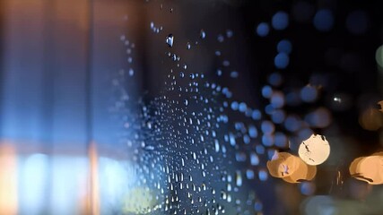 Close-up of rain droplets trickling down a glass window, with a blurred image and soft lights in the background, creating a calm and moody aesthetic.