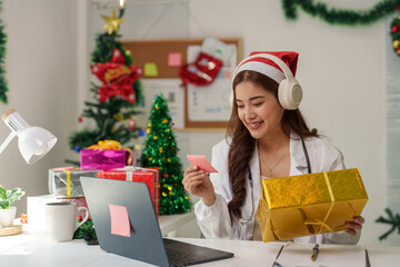 Wall Mural - A female doctor wearing a Santa hat wearing music headphones and holding a gift box is making an online video call from the office. Filled with Christmas decorations Creating a holiday atmosphere.