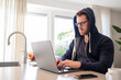 © ReLein - Working at home in the kitchen. caucasian male sitting at the kitchen counter behind his laptop working and communicating. Dressed in a blue hoodie, wearing glasses.
